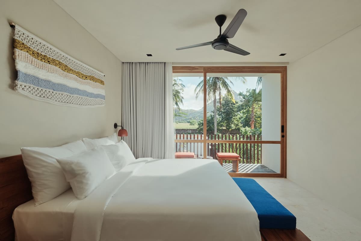 Une chambre avec un lit, un ventilateur de plafond, une vue sur un balcon doté d’une balustrade en bois, et une fenêtre donnant sur des palmiers et des montagnes.