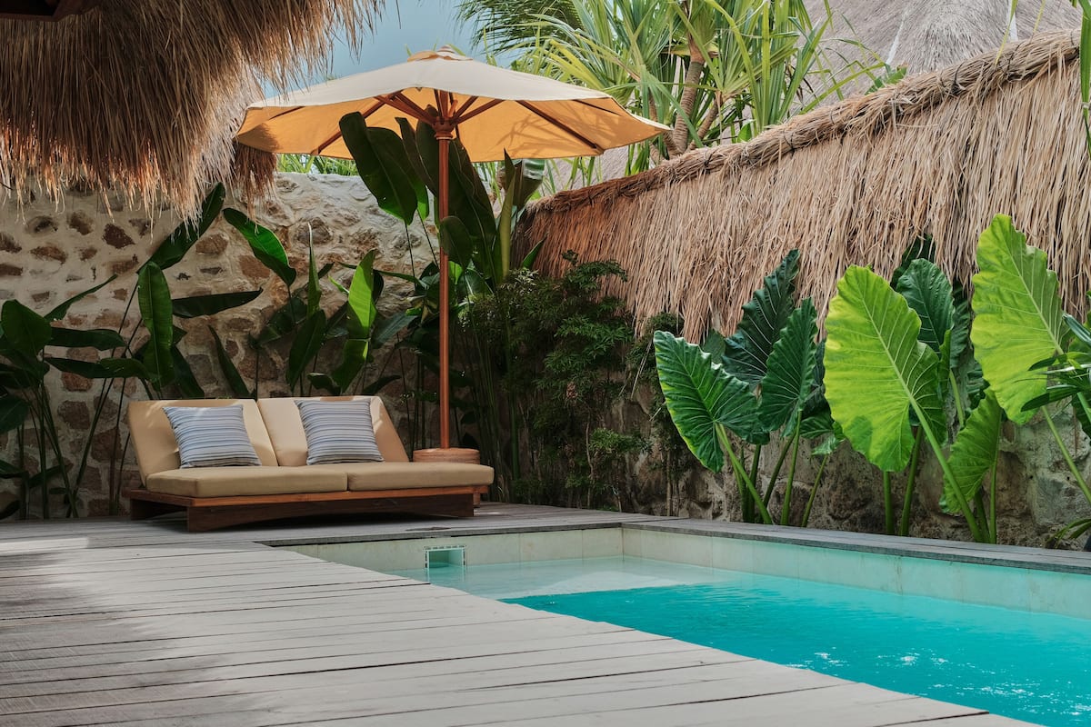 Un espace aménagé au bord de la piscine, avec une terrasse en bois, un canapé beige agrémenté de coussins à rayures, un parasol en paille et des plantes tropicales.