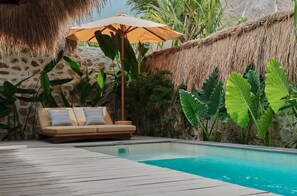 Zona junto a la piscina con terraza de madera, sofá color beige con cojines a rayas, sombrilla de paja y plantas tropicales.