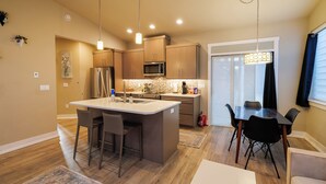 Una cocina moderna con una isla central, taburetes de bar, una mesa de comedor con sillas y una lámpara colgante.