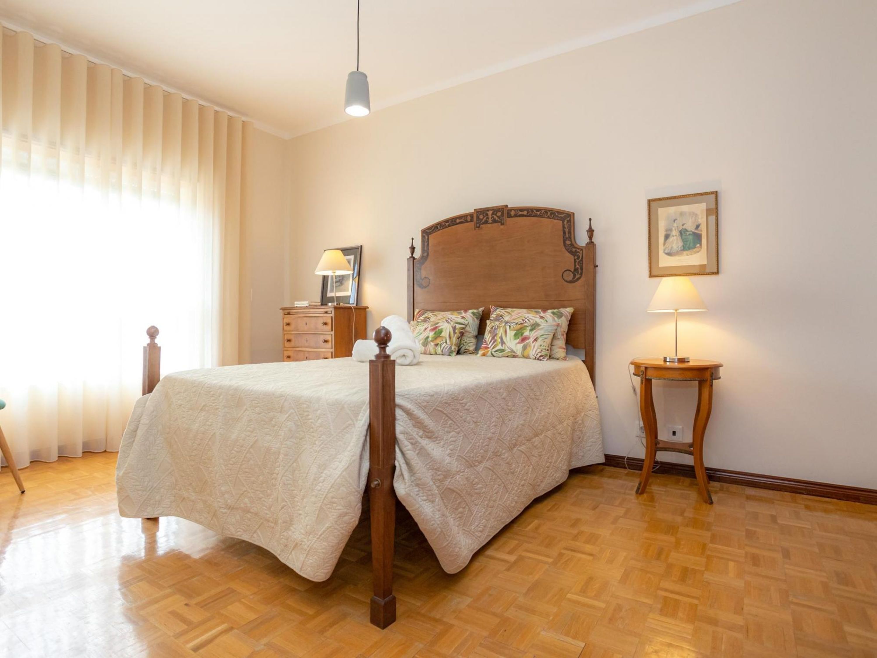 A neatly arranged bedroom with a wooden bed, a bedside table with a lamp, a dresser, and a window with curtains.