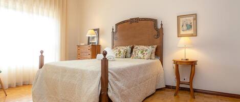 A neatly arranged bedroom with a wooden bed, a bedside table with a lamp, a dresser, and a window with curtains.