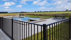 A rectangular swimming pool with a wooden deck and benches, surrounded by a black metal fence. - Large modern holiday home with pool close to the beach (Groede)