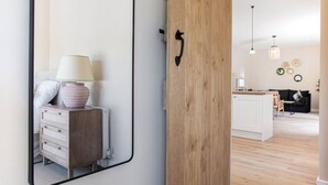 A modern interior with a wooden door, a mirror reflecting a room with a dresser, lamp, and a bed. - The Barn in Longhope - Luxury Barn Conversion (Longhope)
