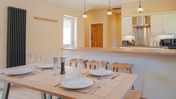 A modern kitchen with wooden countertops, a stainless steel range hood, and a dining area set with plates and glasses. - The Barn in Longhope - Luxury Barn Conversion (Longhope)