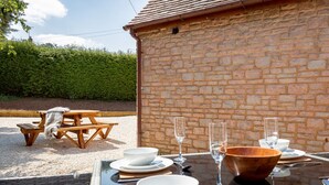 An outdoor dining area with a brick wall, wooden picnic table, and a table set for two with white bowls, glasses, and cutlery. - The Barn in Longhope - Luxury Barn Conversion (Longhope)