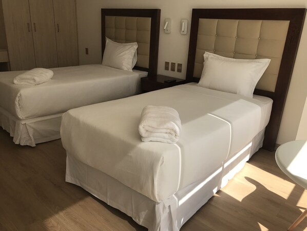 Quarto de hotel com duas camas, cada uma com um edredom branco e uma toalha dobrada. Há mesas de cabeceira com abajures e um guarda-roupa ao fundo.