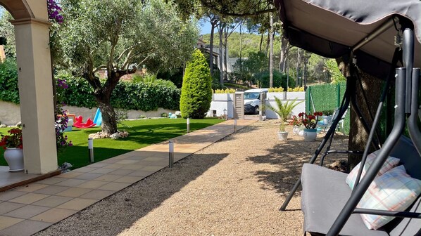 Un espace extérieur aménagé, abrité et doté d’un chemin de gravier, de plantes en pot, et offrant une vue sur une voiture blanche et sur la verdure en arrière-plan.