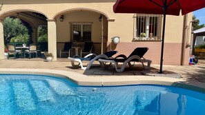 Une piscine avec des chaises longues et un parasol rouge, un patio avec des chaises et une table, et un bâtiment doté d’arcades et d’une fenêtre ornée de plantes.