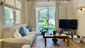 Un salon avec un canapé, une télévision et une vue sur un jardin, visible à travers une porte vitrée.