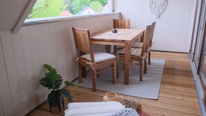 Una mesa de comedor de madera con cuatro sillas, una planta pequeña y un tapiz de macramé en una habitación con piso de madera y una ventana con vista a la vegetación.