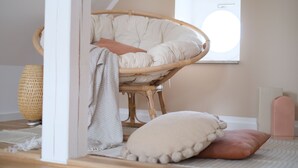 Una habitación con una silla de ratán, un ventanal grande, una cesta tejida y una cama con almohadas.