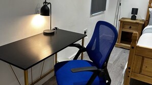 Um quarto pequeno e bem iluminado, com uma mesa, uma cadeira de escritório azul, uma mesinha de cabeceira de madeira e uma cama.