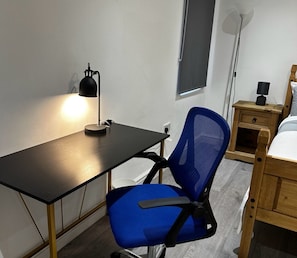 Une petite pièce bien éclairée, avec un bureau, une chaise de bureau bleue, une table de chevet en bois et un lit.