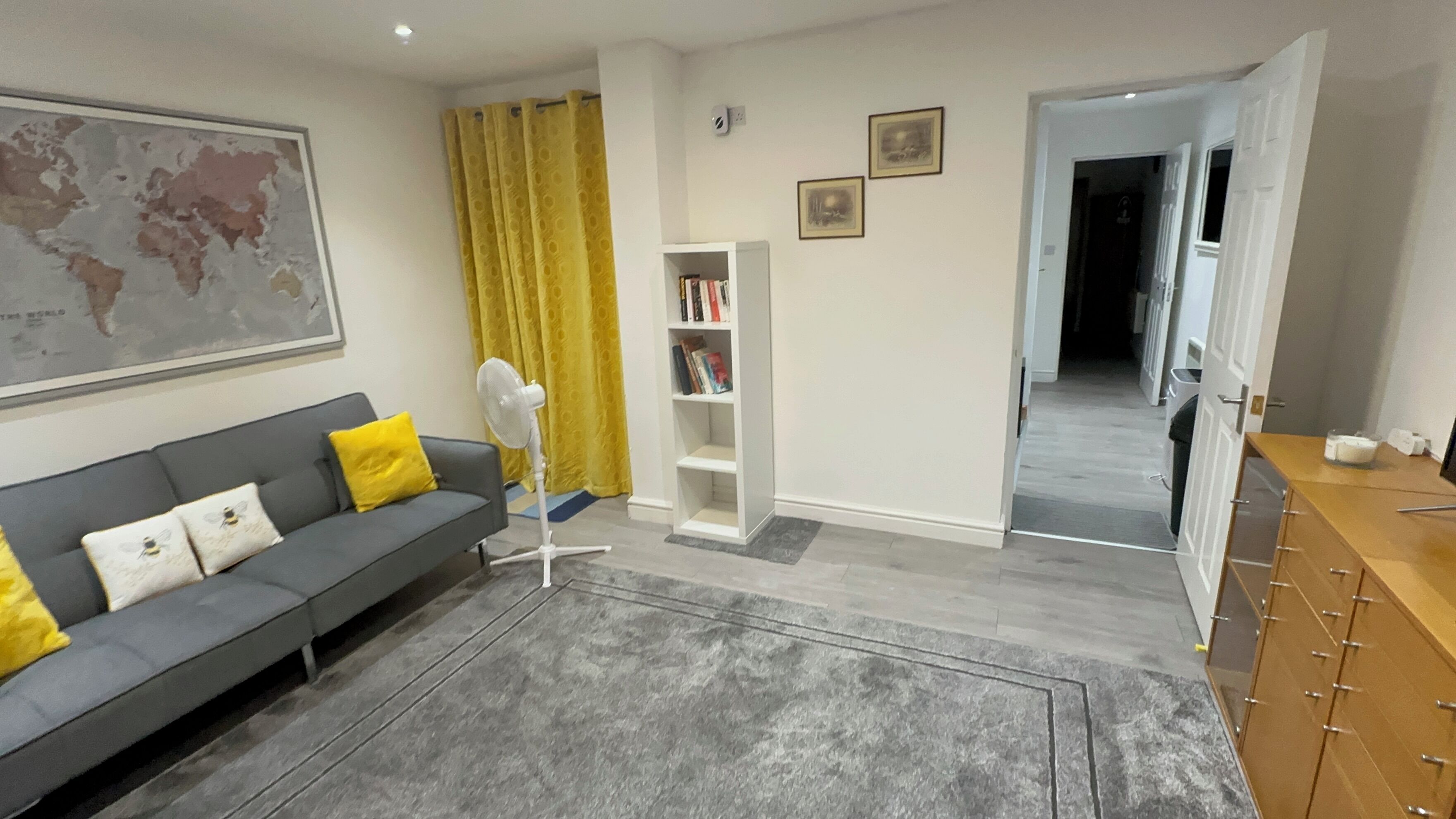 A modern living room with a grey sofa, a standing fan, a bookshelf, and a world map on the wall.