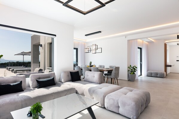 A modern living room with a sofa, ottoman, and a glass coffee table. There is a dining area with chairs and a table in the background.