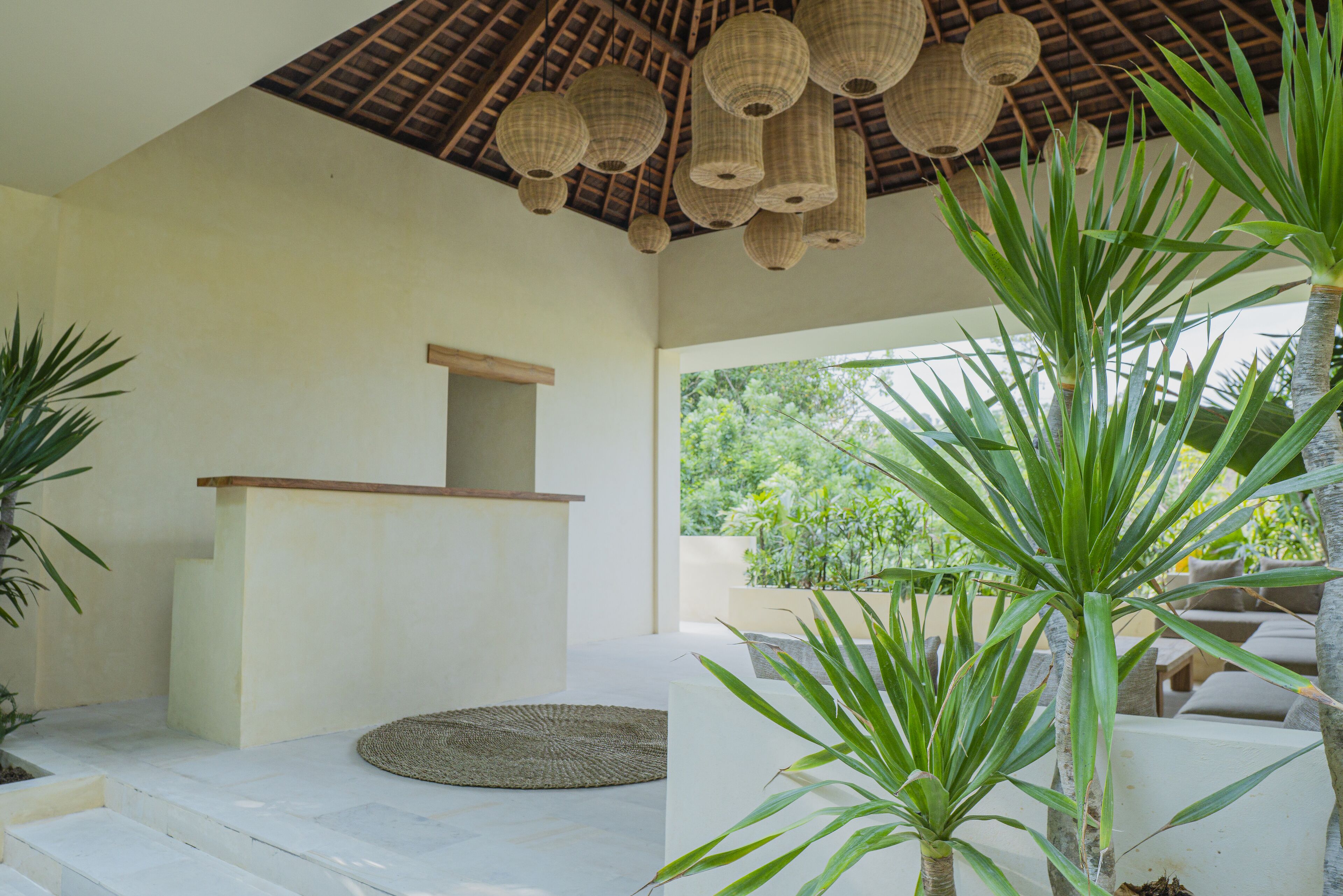 A modern interior with a wooden ceiling, woven light fixtures, a reception desk, and a view of greenery outside.