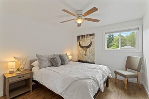 Une chambre à coucher comprenant un lit, une table de chevet, une chaise, un ventilateur de plafond et une fenêtre donnant sur des arbres.