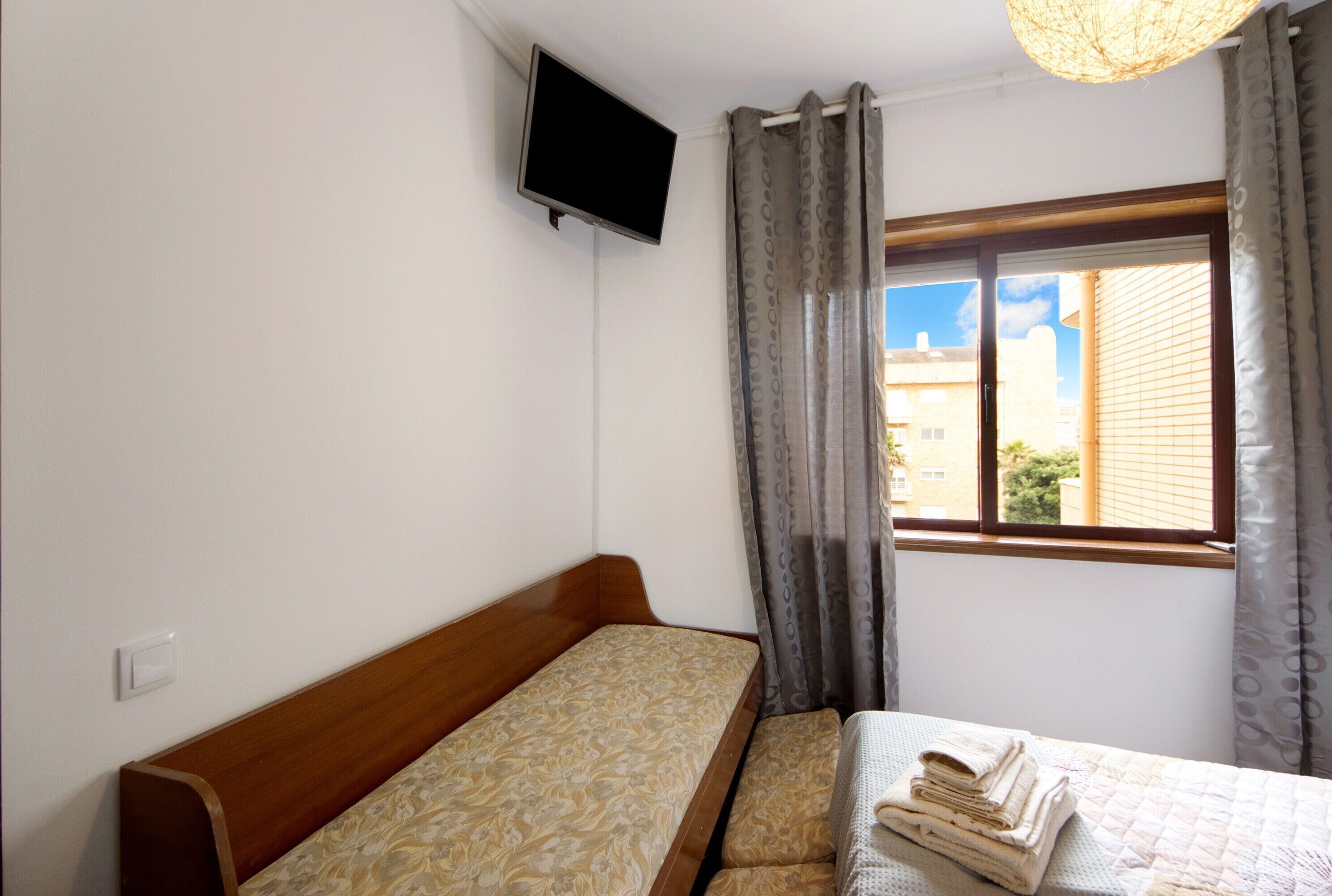 A single bed with a wooden headboard, a flat-screen TV mounted on the wall, and a window with curtains.