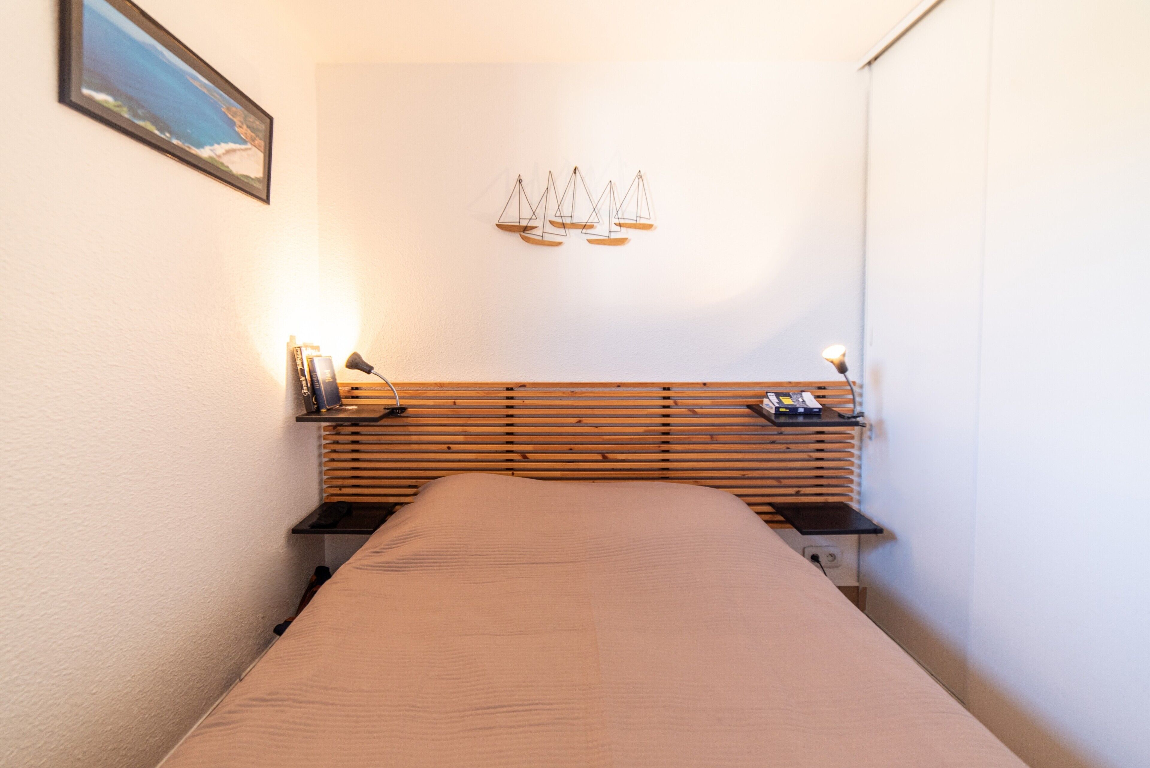 A narrow hotel room with a wooden headboard, a bed, a small desk with a lamp, and a wall-mounted light fixture.