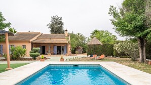 Ein Pool mit einer Fliesenbordüre, ein Haus mit einem Ziegeldach und ein Sonnenschirm mit Reetdach.