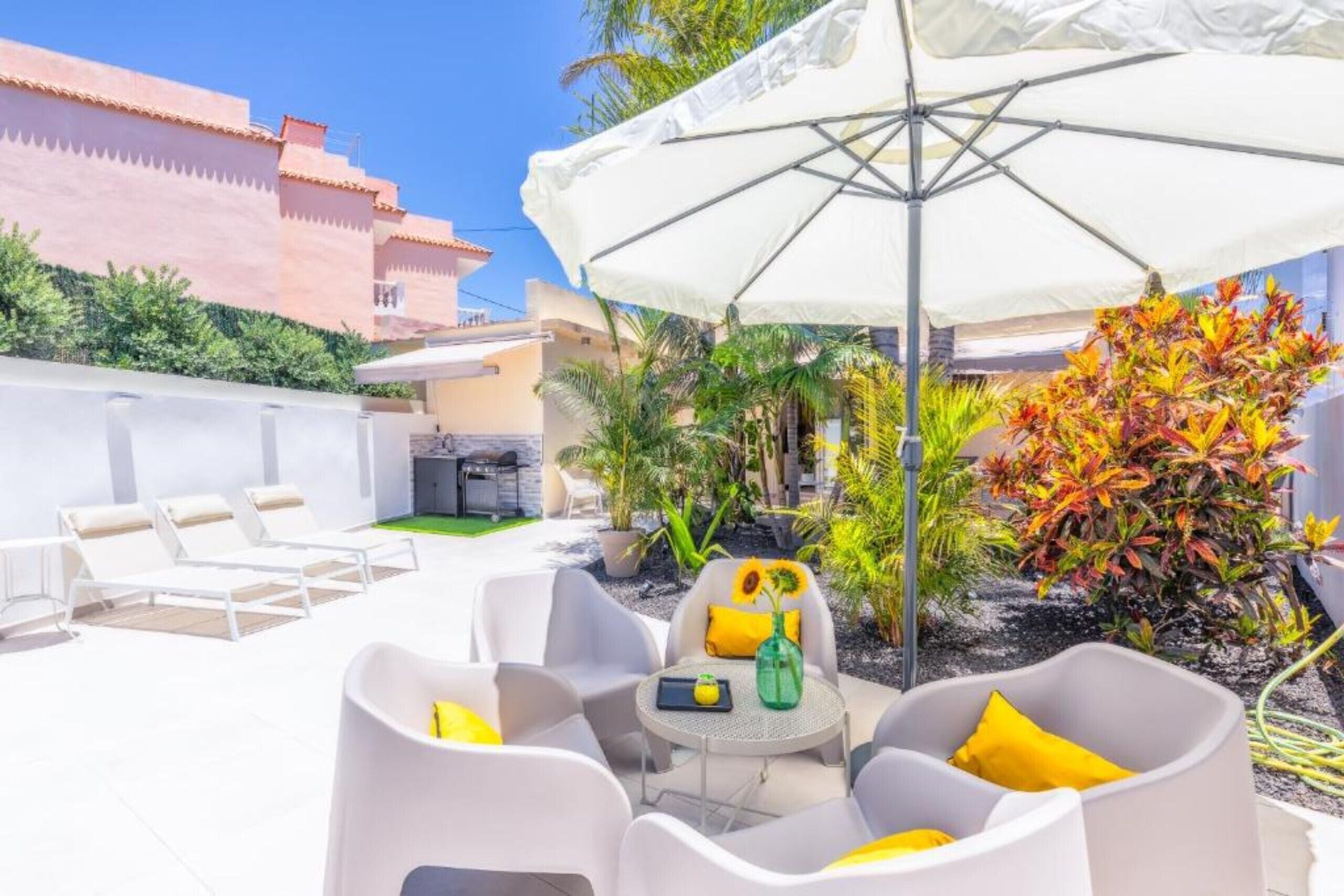Une terrasse avec des meubles blancs, une table agrémentée d’un vase et d’un plateau, et un grand parasol.