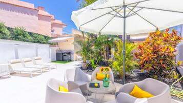 Une terrasse avec des meubles blancs, une table agrémentée d’un vase et d’un plateau, et un grand parasol.