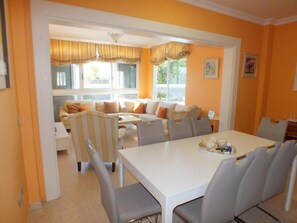 Un coin repas avec une table blanche et des chaises grises, un canapé avec des coussins orange et une fenêtre avec des rideaux.