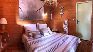 A wooden cabin bedroom with a bed, a nightstand, a lamp, a chair, a wall-mounted shelf with a plant, a hanging light fixture, and a door. - Gîte Escale Bien Etre in Biscarrosse (Biscarrosse)