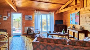 A wooden cabin interior with a sofa, dining table, chairs, a television, and a sliding glass door leading to an outdoor area. - Gîte Escale Bien Etre in Biscarrosse (Biscarrosse)