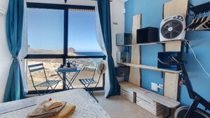 A room with a bed, a table with towels, a chair, a fan, and a view of the beach.