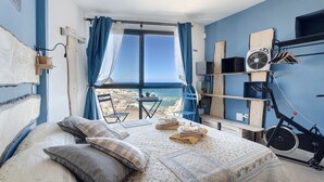 A bedroom with a bed, pillows, a window with curtains, a fan, and a view of the sea.
