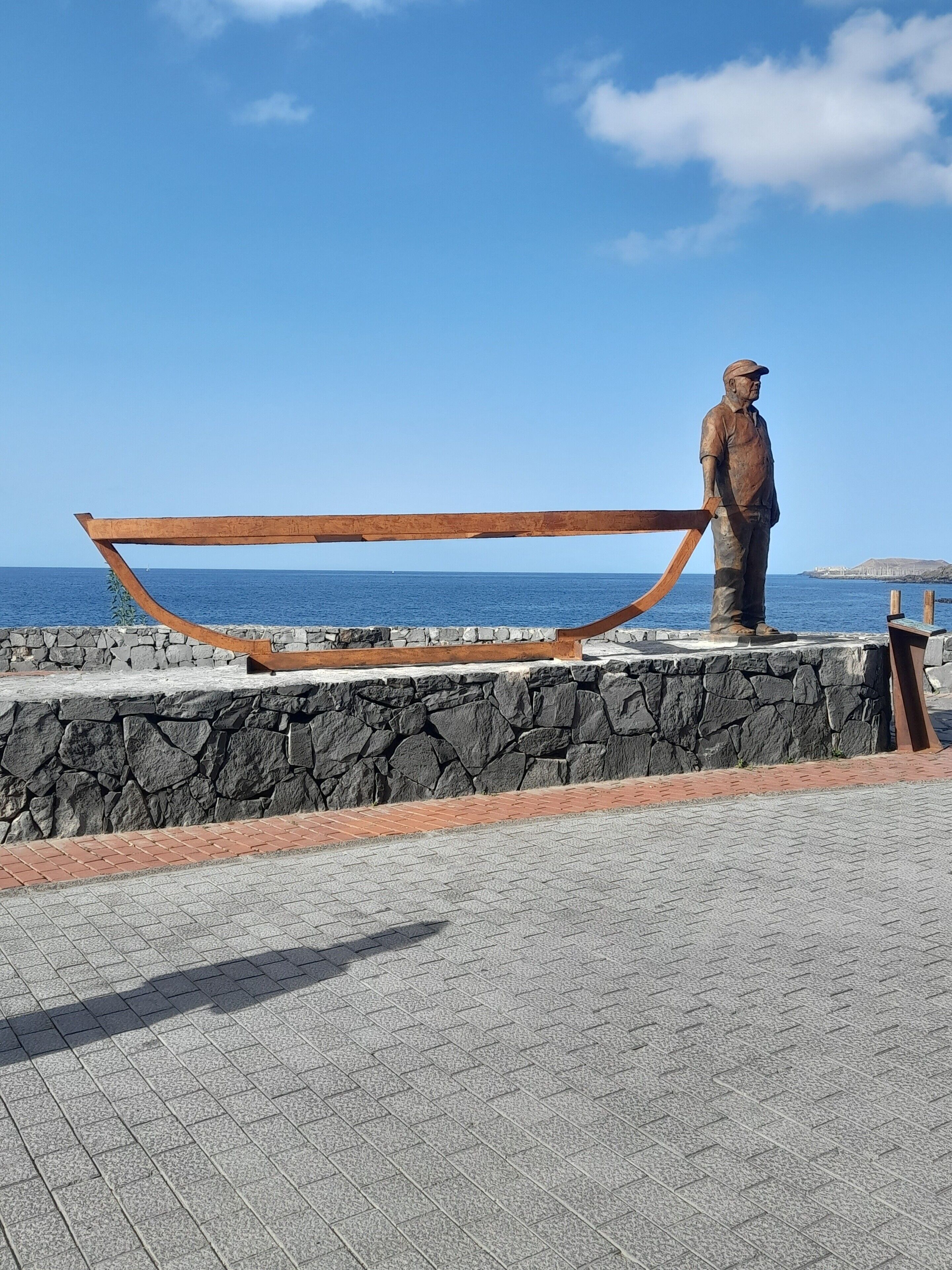 A statue of a man holding a boat on a stone wall by the sea.