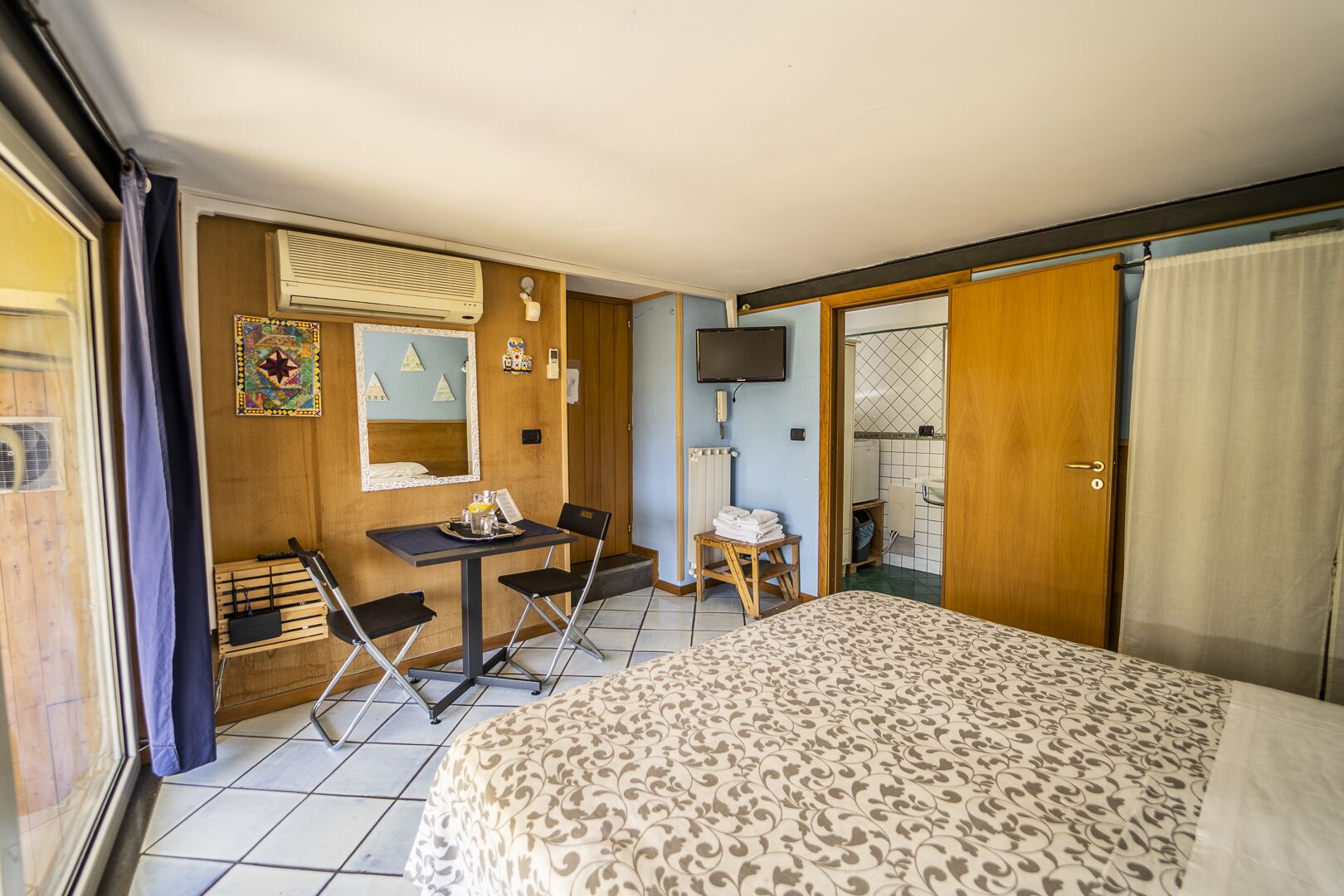 A hotel room with a bed, a desk with chairs, a television, and a bathroom visible through an open door.