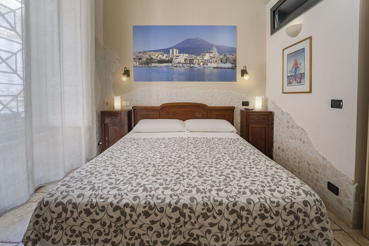 A bedroom with a bed, two bedside tables, a nightstand, a wall-mounted lamp, a framed picture, and a window with curtains.