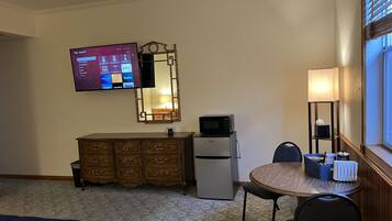 A hotel room with a TV mounted on the wall, a microwave on a mini-fridge, a wooden dresser, a round dining table with a chair, and a lamp.