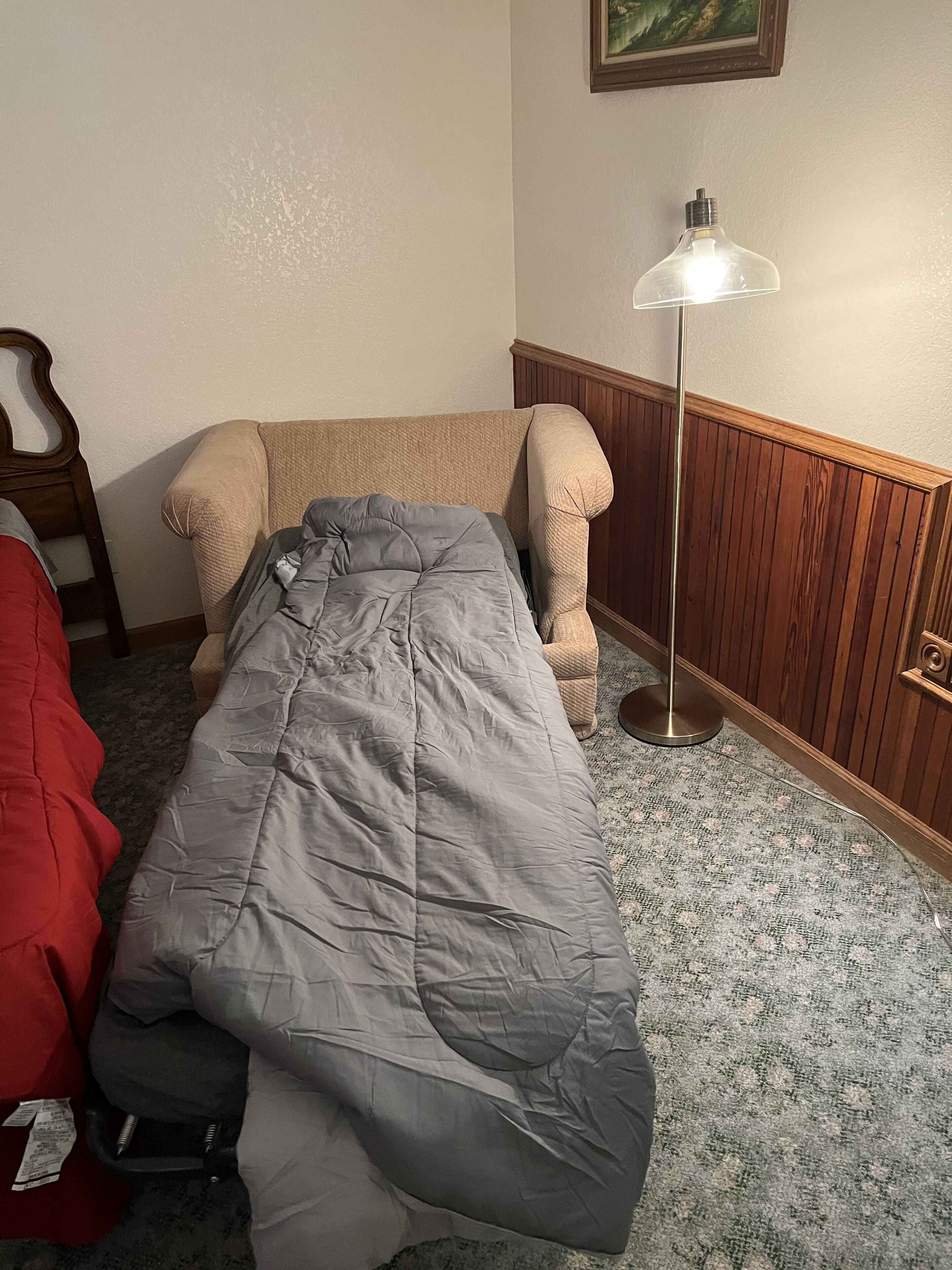A single bed with a grey blanket, a wooden headboard, a red blanket, a floor lamp, and a patterned carpet.