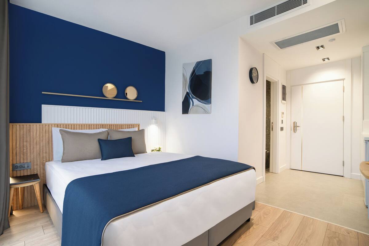 A modern bedroom with a large bed, a bedside table, a nightstand, a wall-mounted shelf with decorative items, a painting, and a door leading to another room.