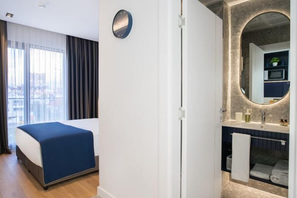 A modern hotel room with a bed, a large window with curtains, a bathroom with a mirror, sink, and towel rack.