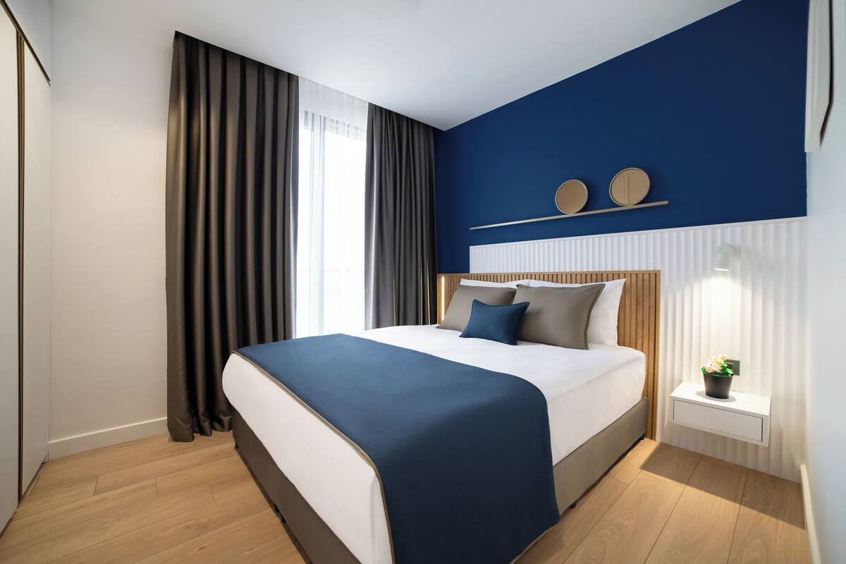 A modern hotel room with a large bed, a bedside table with a lamp, and a window with curtains.