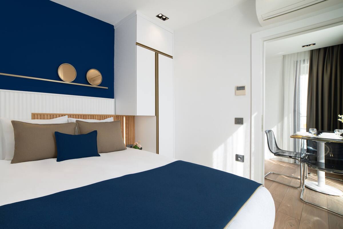 A modern hotel room with a large bed, a wooden headboard, a blue accent wall, and a dining area with a glass table and chairs.