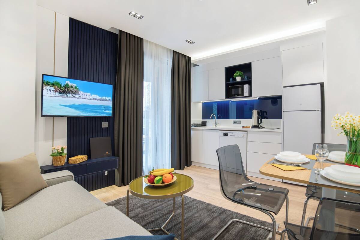 A modern hotel room with a flat-screen TV, a dining area with a table set for two, and a kitchenette.