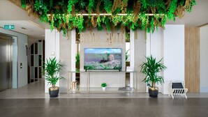 Un área de recepción con una pantalla de televisión mostrando una escena natural, un cartel que dice “Still Suite Marine Hotel” y dos plantas en macetas.