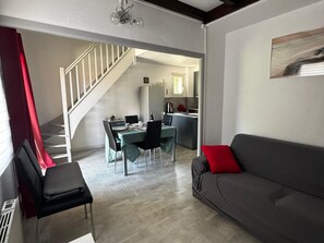A modern living room with a grey sofa, a dining area with a table and chairs, and a kitchen in the background.