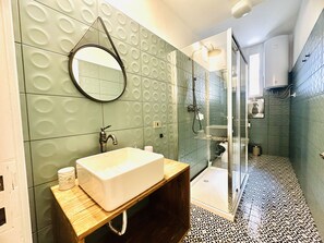 A bathroom with a white sink, a wooden vanity, a round mirror, and a shower area with a glass door. - Historical building in the heart of the city center  (Napoli)
