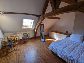 Quarto com piso de madeira, cama com roupa de cama branca, uma mesinha com cadeiras, uma janela e teto inclinado.