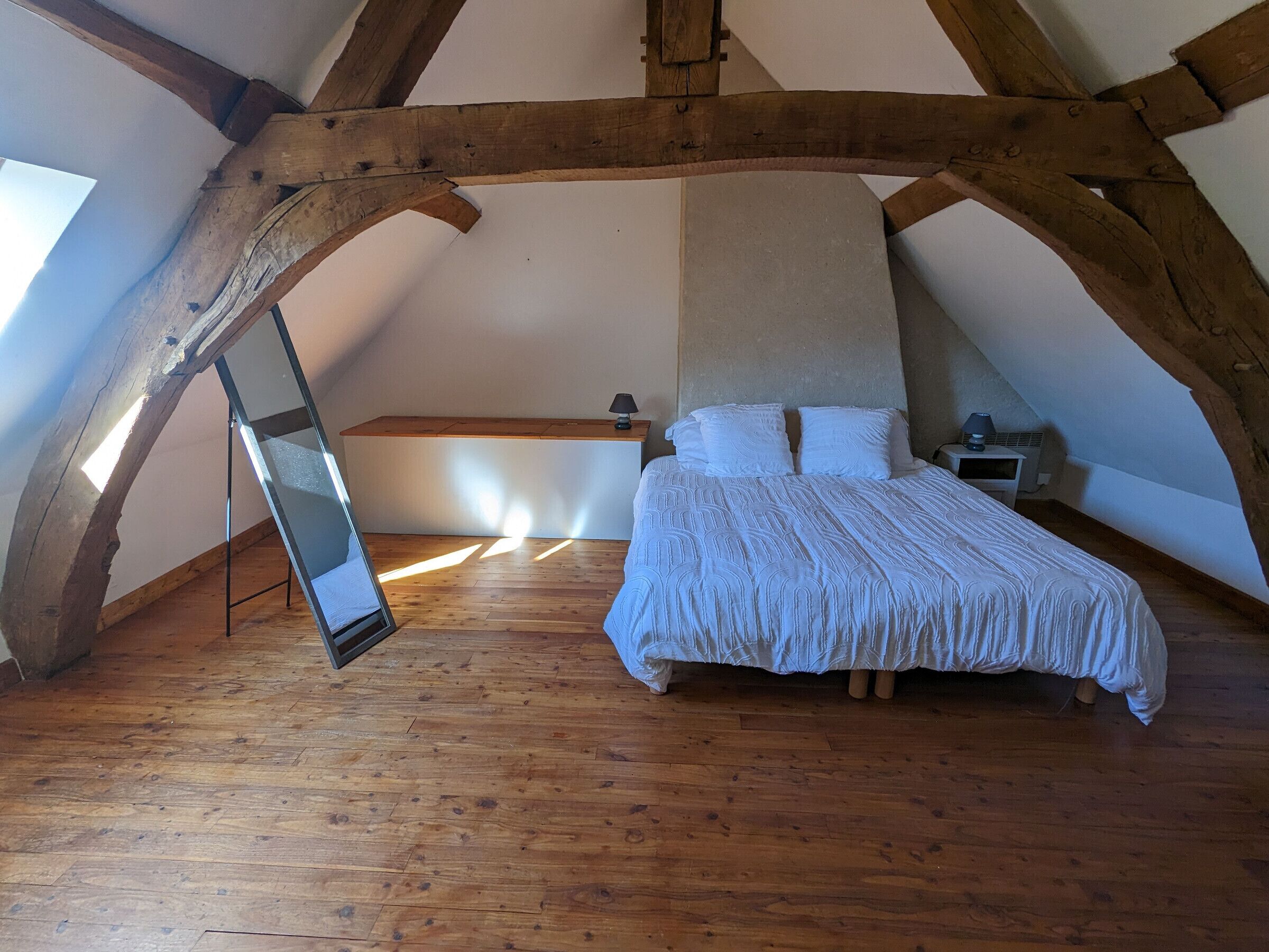 A bedroom with a sloped ceiling, wooden beams, a bed with white bedding, a bedside table with a lamp, and a mirror on the wall.