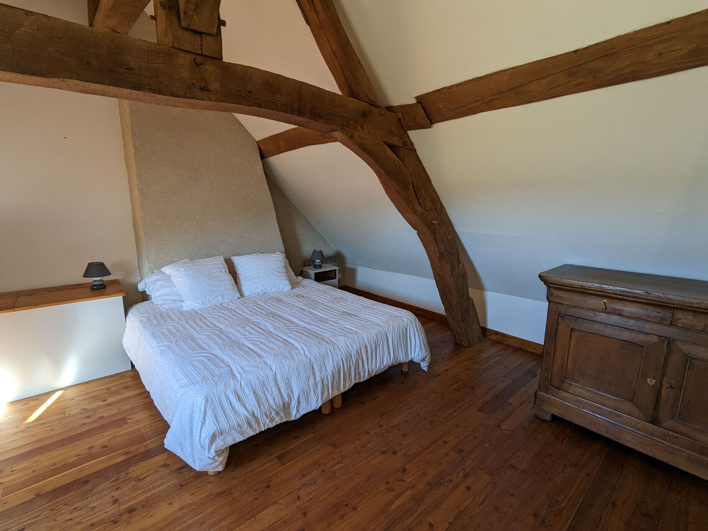 Een slaapkamer met een houten plafond, een bed met wit beddengoed, een nachtkastje met een lamp en een houten dressoir.