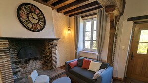 A cozy living room with a stone fireplace, a blue sofa with cushions, two white chairs, and a large wall clock.