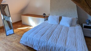A bedroom with a wooden ceiling, a bed with white bedding, a bedside table with a lamp, and a small white cabinet.